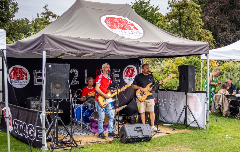 Wide photograph of Craig Hodgson performing with the full band on the EH32 Live outdoor stage beneath the event canopy.